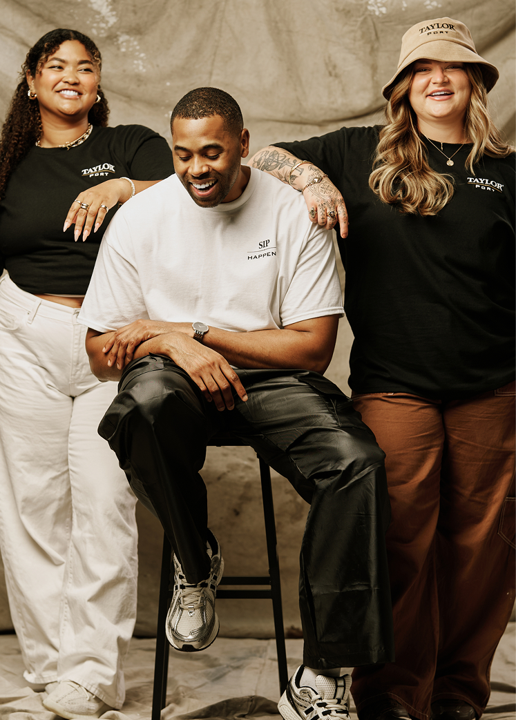 Man sitting on the stool with two girls on each side wearing Taylor Port Merch